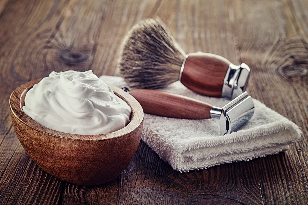 Wooden bowl with shaving cream, a shaving brush, and razor on a white towel on wooden surface.